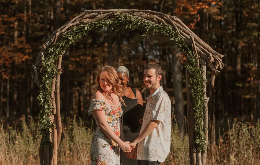 A couple in a floral dress and a cream shirt hold hands under a branch arch in an autumn field while the officiant stands between them