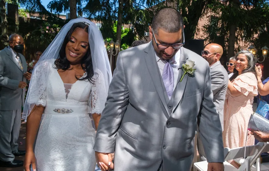 A bride and groom walk back up the aisle hand in hand past a small group of standing guests in a leafy courtyard