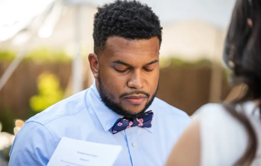 A man in a blue shirt and bow tie looks down at a printed page he is reading aloud at an outdoor wedding
