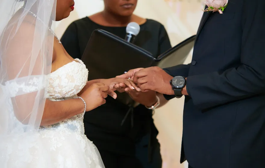A groom slides a ring onto his bride's finger while the officiant holds the microphone and an open binder behind their hands