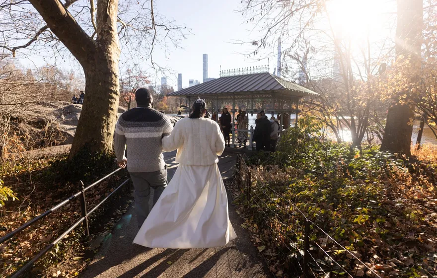 A couple walks arm in arm down the paved path toward the Ladies' Pavilion on a sunny autumn day, trees on both sides