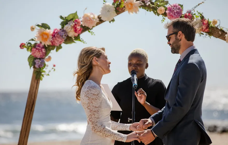 An officiant speaks into a microphone on a stand with her notes in hand while a couple holds hands under a floral arch on a beach