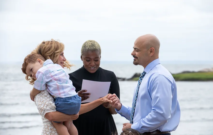 A bride holds a toddler on her hip while the groom takes her hand beside the officiant on a beach