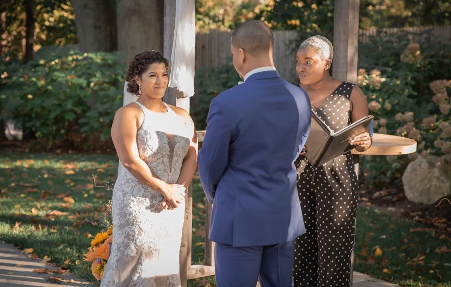 An officiant in a polka-dot jumpsuit reads from an open black binder while a bride and groom stand facing her in a backyard