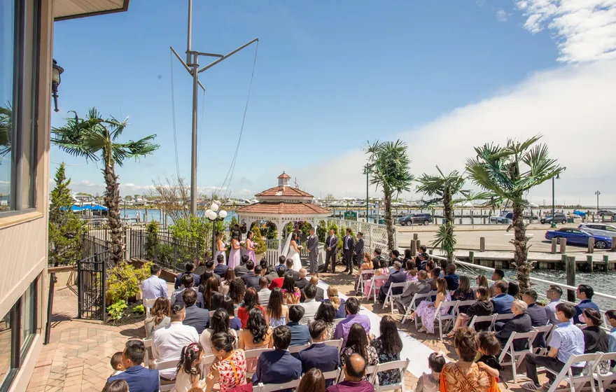 Rows of seated guests at a waterfront ceremony watch the couple and officiant under a white gazebo, palm trees and blue sky above