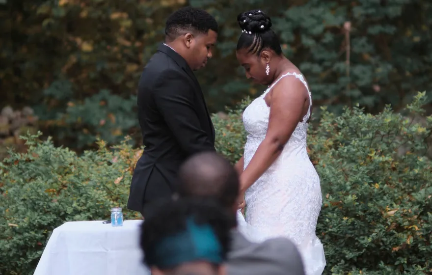 A bride and groom stand with heads bowed at a white-clothed table that holds a jar of sand, guests in the foreground