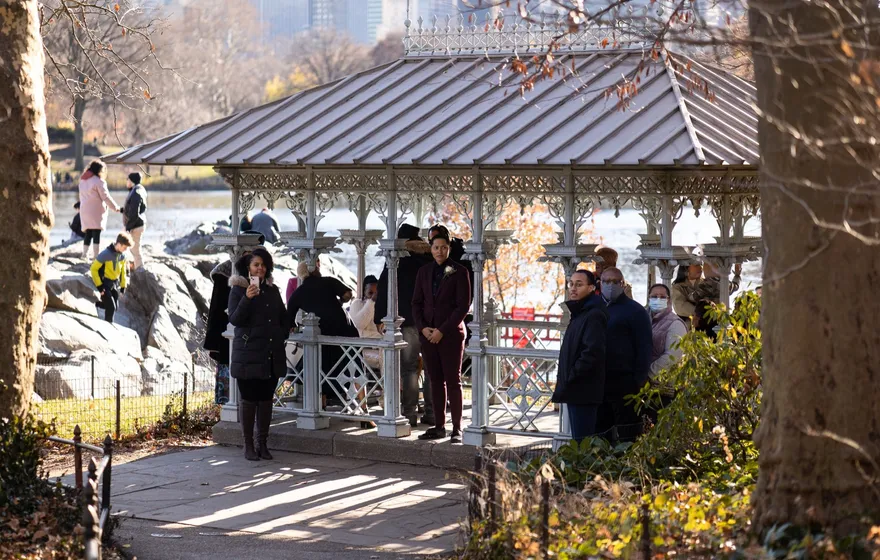 The Ladies' Pavilion seen from the path, a wedding party gathered inside and guests standing on the paving outside, with the Lake and city buildings behind