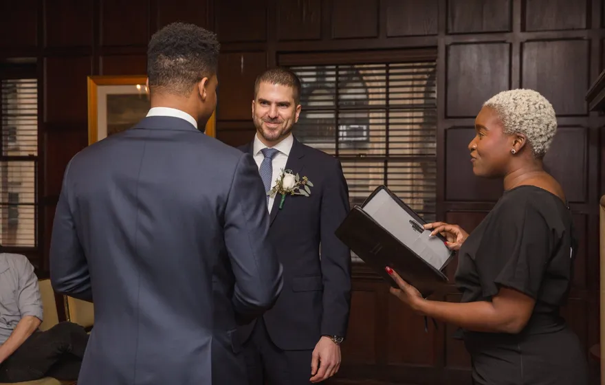 In a wood-paneled room, the officiant reads from an open folder to two grooms in navy suits, one smiling at her