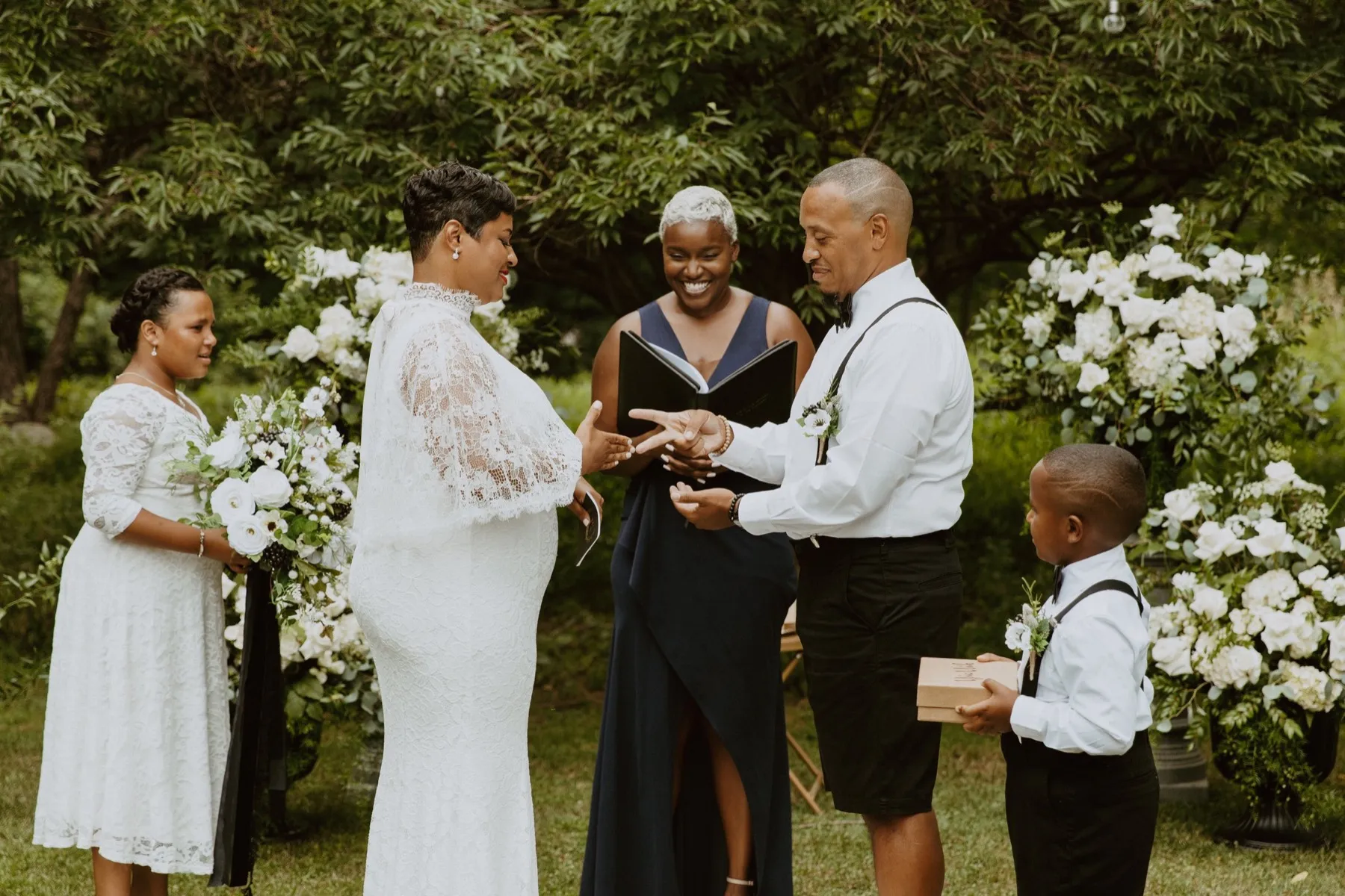 A couple exchanges rings during a garden vow renewal as the officiant smiles between them and two family members stand alongside