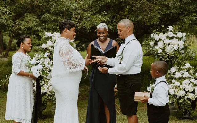 A couple exchanges rings during a garden vow renewal as the officiant smiles between them and two family members stand alongside