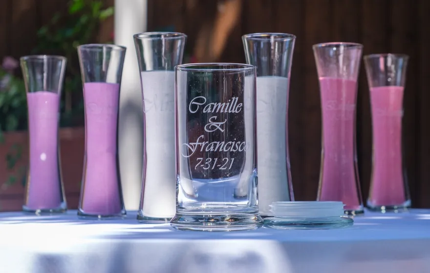 Six slim glass vases of purple, white and pink sand arranged around a larger engraved glass vessel on a white tablecloth