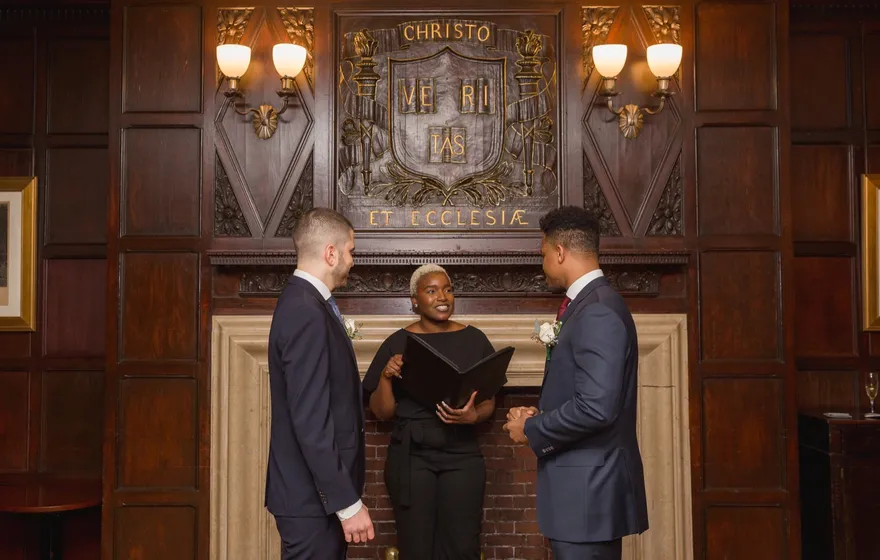 Two grooms in navy suits stand before the officiant at a carved wooden fireplace in a wood-paneled club room