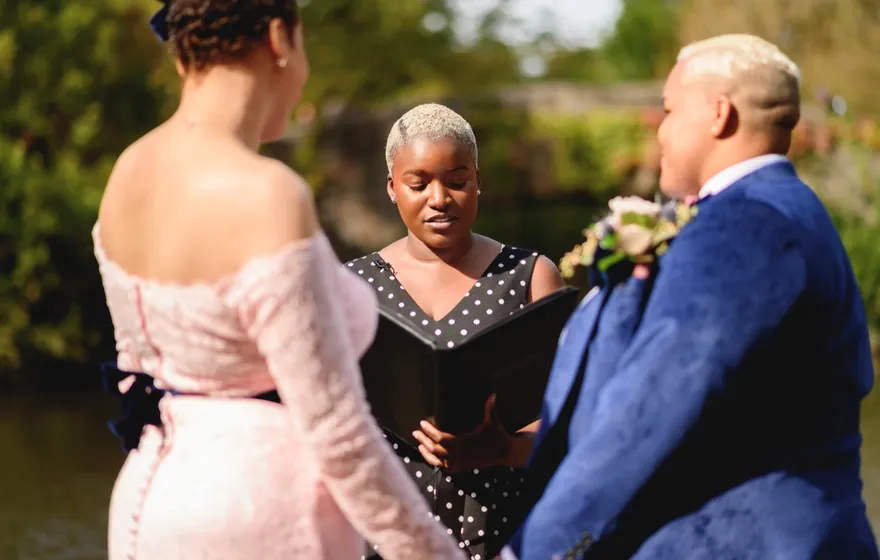 An officiant reads from an open black binder while standing between a couple beside the water in Central Park