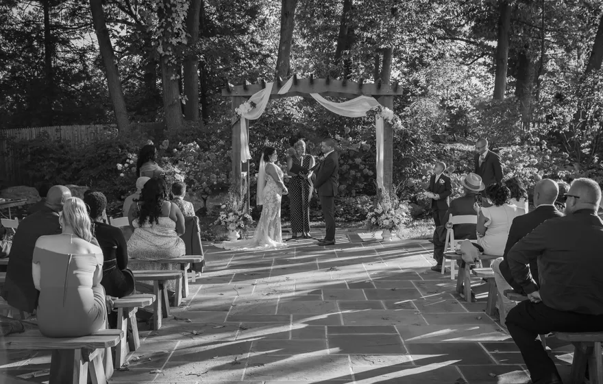 Black and white view from the back of a garden aisle, guests on wooden benches on both sides and the couple with the officiant under a draped pergola at the far end