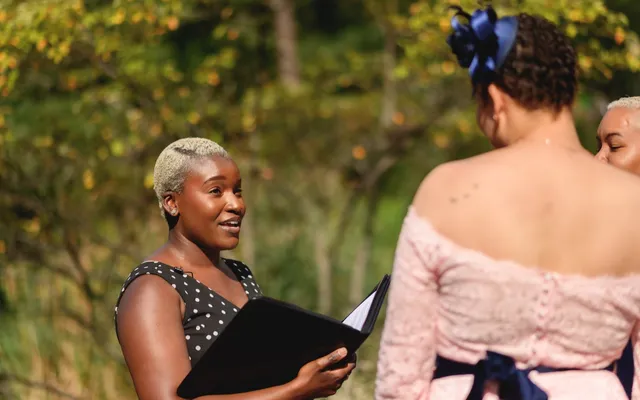 An officiant with an open ceremony binder speaks to a couple during an outdoor vow renewal