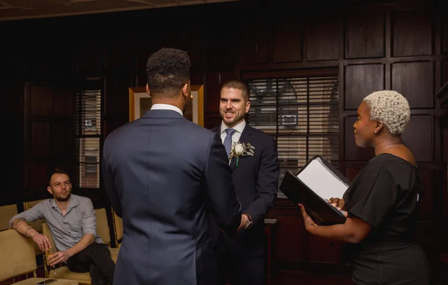 Two grooms face each other in a wood-paneled club room while the officiant reads from a binder and a single guest watches from an armchair