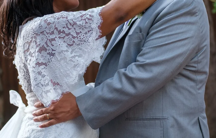 A bride in a lace dress wraps her arm around her groom's neck as he holds her waist, the two looking at each other