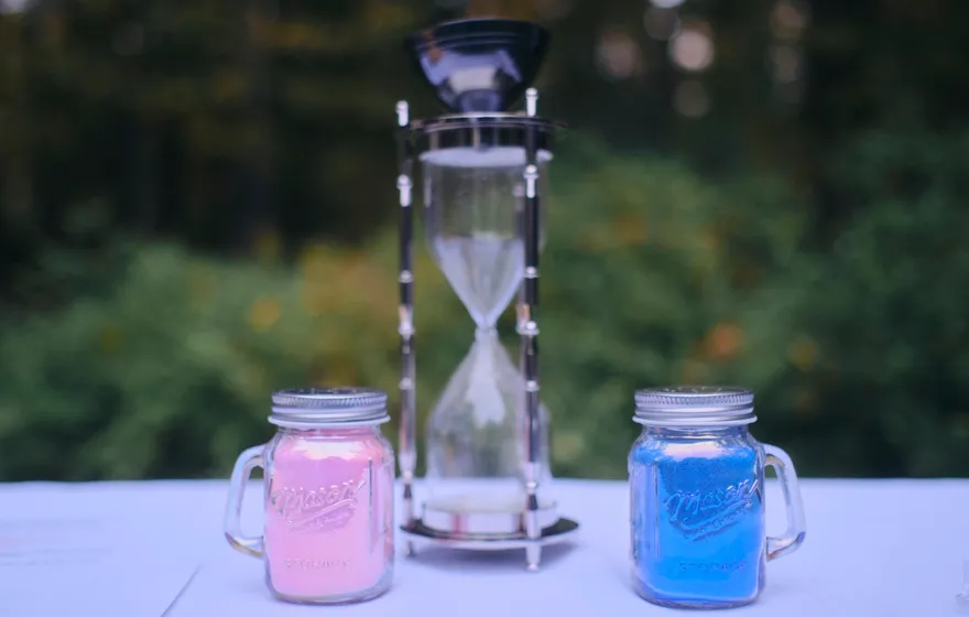 Two small mason jars of pink and blue sand sit in front of an hourglass on an outdoor table