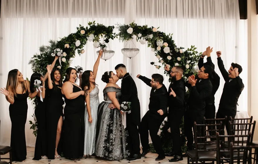Under a tall floral arch, a couple dressed in black kisses while their wedding party throws their arms up and cheers on both sides