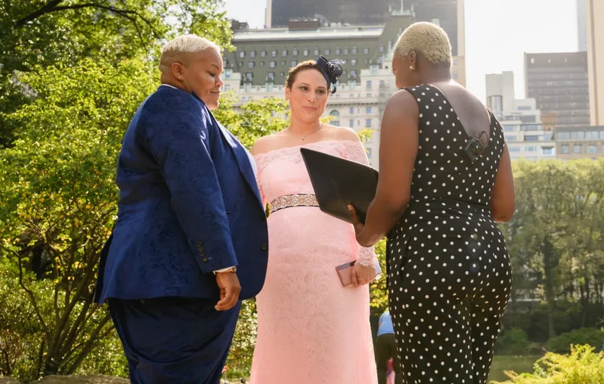 The officiant holds an open binder while a couple in a blue suit and a pink lace gown stand on a Central Park path with tall buildings behind the trees