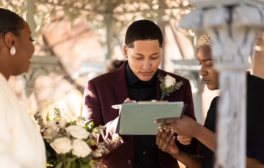 A newlywed in a burgundy suit signs on a clipboard the officiant holds steady, a bouquet of white roses in the foreground