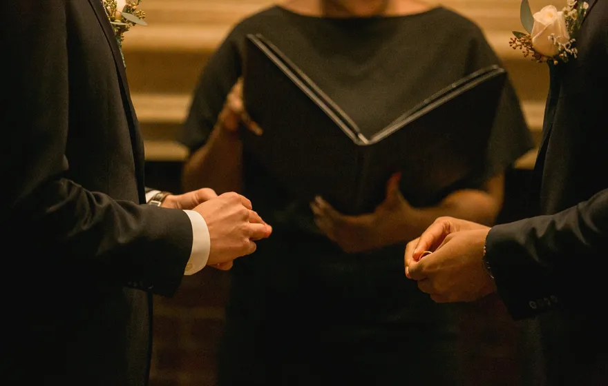 Two grooms in dark suits each hold a ring between their fingers while the officiant stands between them with an open binder