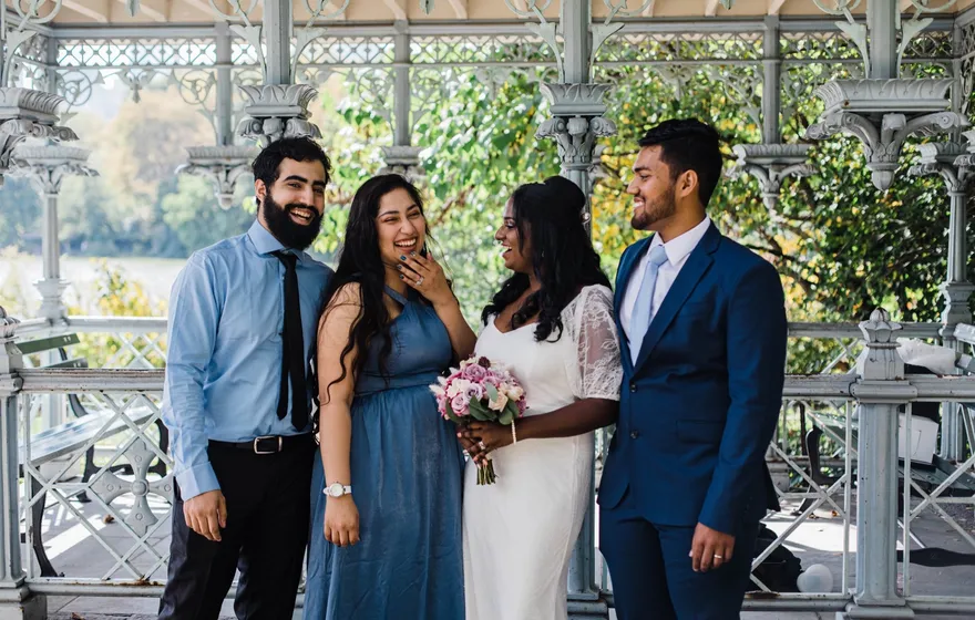 Two couples stand together inside the white cast-iron Ladies' Pavilion in Central Park, the Lake visible through the ironwork behind them