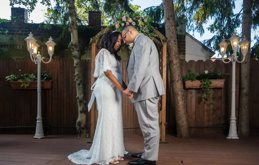 A bride and groom hold hands under a flowered wooden arch in a small brick-walled courtyard strung with lantern lamps