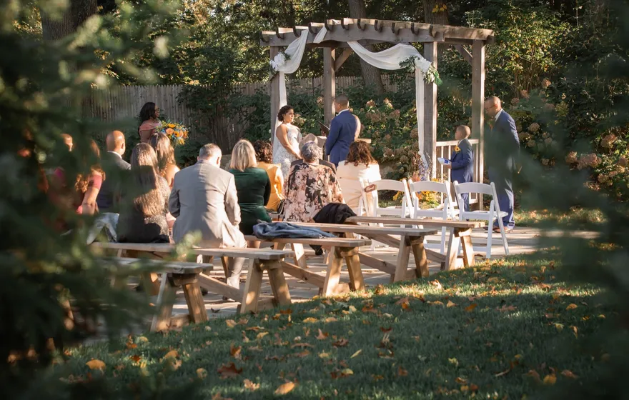 A garden ceremony seen through the trees, guests seated on wooden benches on the lawn and the couple standing under a draped wooden arbor