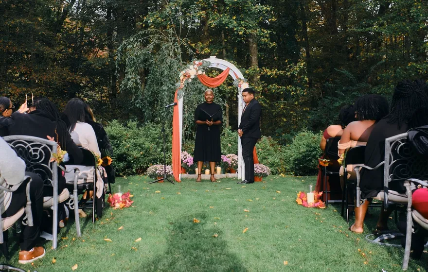 An officiant in black speaks at a white arch on a lawn while the groom waits beside her and seated guests watch from both sides of the aisle