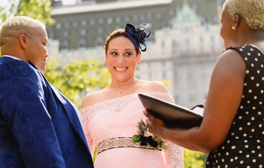 A spouse in a pink lace dress smiles at her partner during a vow renewal while the officiant holds an open binder, Manhattan buildings behind the trees