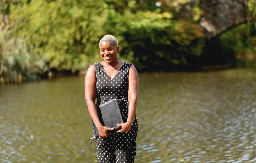 Robyn stands by the water holding a black ceremony binder against her side, smiling