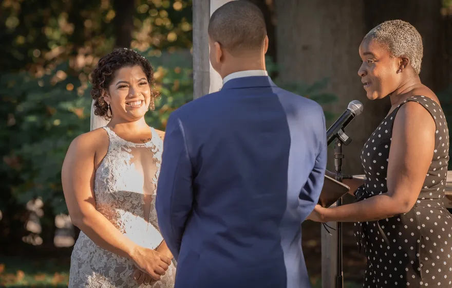An officiant speaks into a microphone on a stand, facing a bride who smiles at her groom