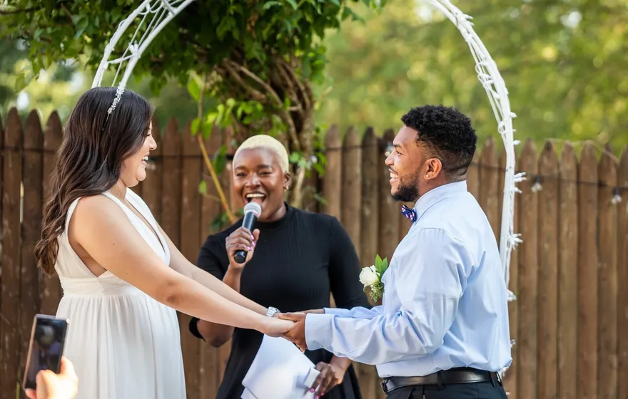 An officiant laughs into a handheld microphone while a couple holds hands in front of her under a white wire arch
