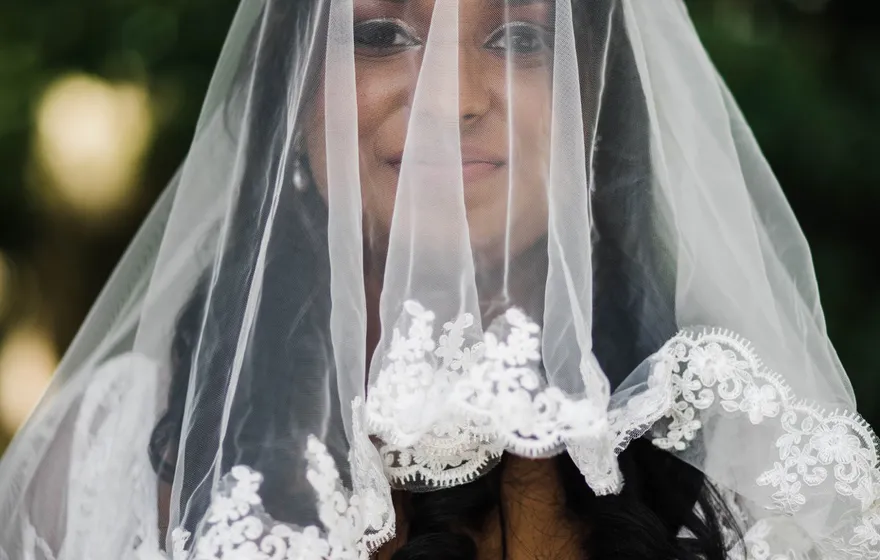 A bride looks straight at the camera through a sheer lace-edged veil that covers her face, holding pink roses