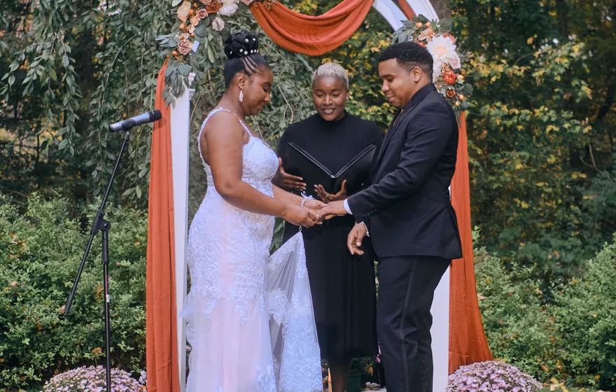 A bride and groom hold hands under a white garden arch draped in rust fabric while the officiant stands between them with an open black binder