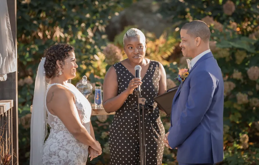 An officiant speaks into a microphone on a stand, looking at the bride, while the groom smiles beside her in a garden