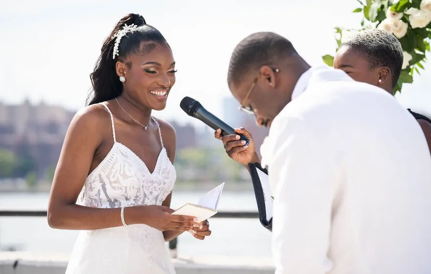 A bride smiles down at a folded card of vows while her groom, head bowed, wipes his eyes beside the officiant on a rooftop