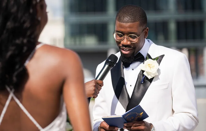 A groom in a white tuxedo jacket and glasses reads his vows from a small booklet while a microphone is held for him
