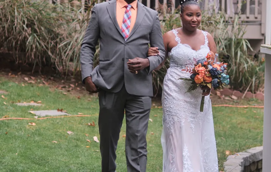 A man in a gray suit and pink tie walks a bride in a lace gown and holding an orange bouquet across a garden lawn