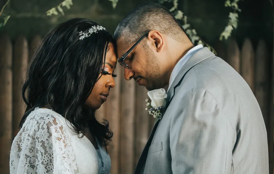 A bride in lace and a groom in a gray suit stand with their foreheads together and their eyes closed