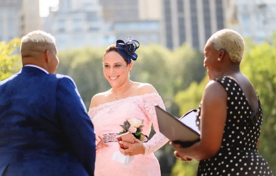 A woman in a pink lace dress smiles at her spouse while the officiant, seen from behind, holds an open binder