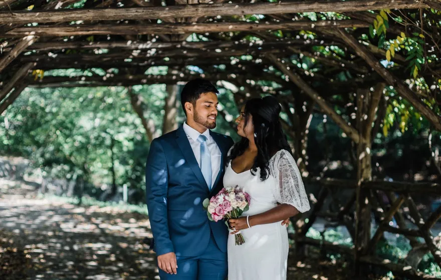 A groom in a blue suit and a bride holding pink roses stand together under a rustic wooden arbor on a shaded Central Park path