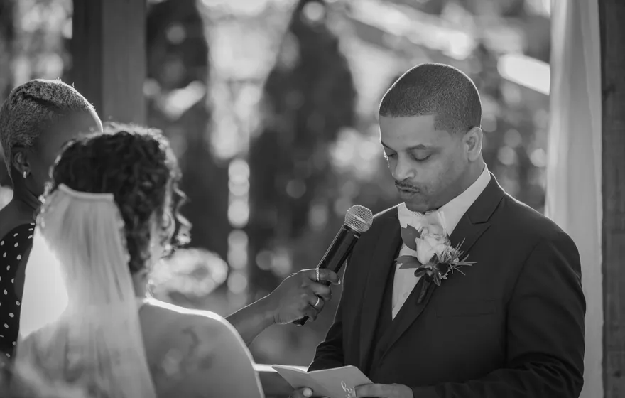 Black and white photo of a man in a suit reading from a card at a microphone during a wedding ceremony