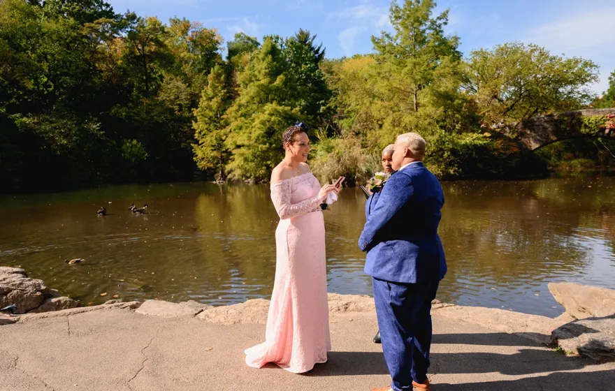 A couple in a pink lace gown and a blue suit face each other on the rocks at the edge of the lake in Central Park, the officiant between them