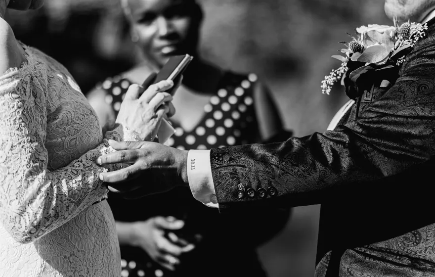 Black and white close-up of one spouse's hand resting on the other's ring hand while the officiant holds a pen and binder behind them