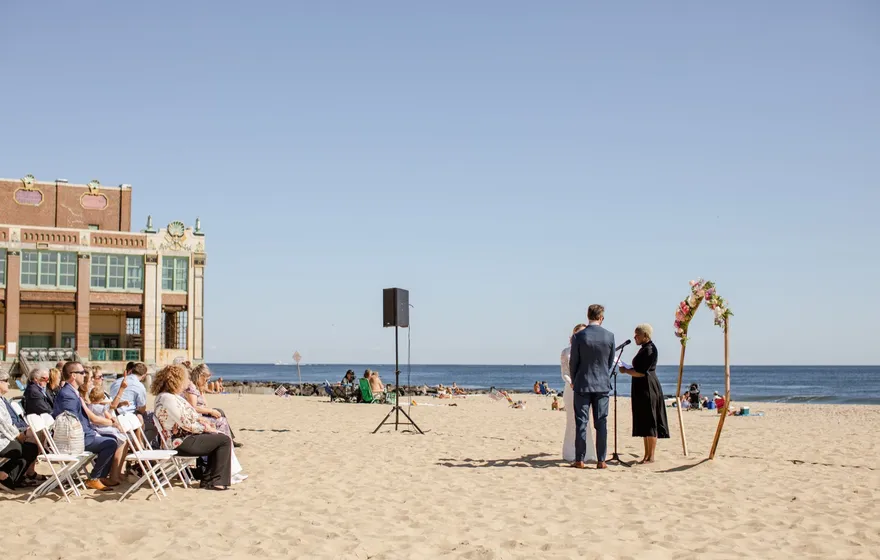 A wide view of a beach wedding with guests seated on the sand, a speaker on a stand and the couple at a wooden arch by the water