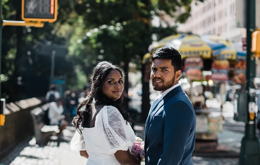 A bride in a white dress with sheer sleeves and a groom in a navy suit stand together at a crosswalk on Central Park South, a hot dog cart behind them