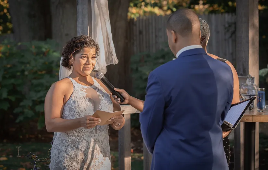 A bride in a lace dress reads her vows from a card to her groom as the officiant holds the microphone for her