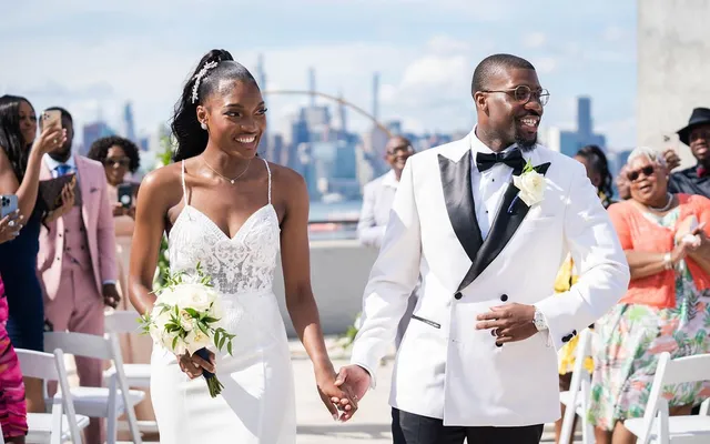 A bride and groom walk back up the aisle hand in hand, smiling, as guests cheer on a rooftop overlooking the city